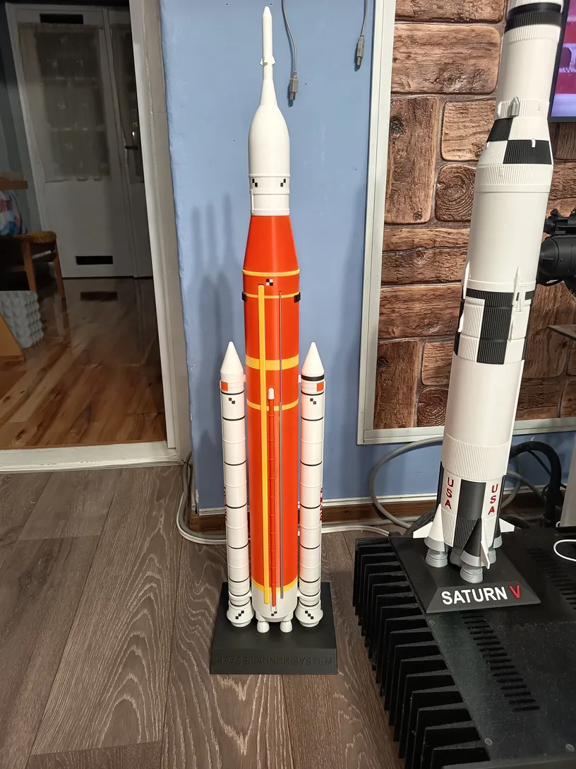 Narancs központi tartályos SLS rakéta makettje a padlón, mellette a fehér Saturn V talapzaton.